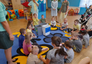 Przedszkolaki siedząc na dywanie tworzą koło, w środku kosze na śmieci.